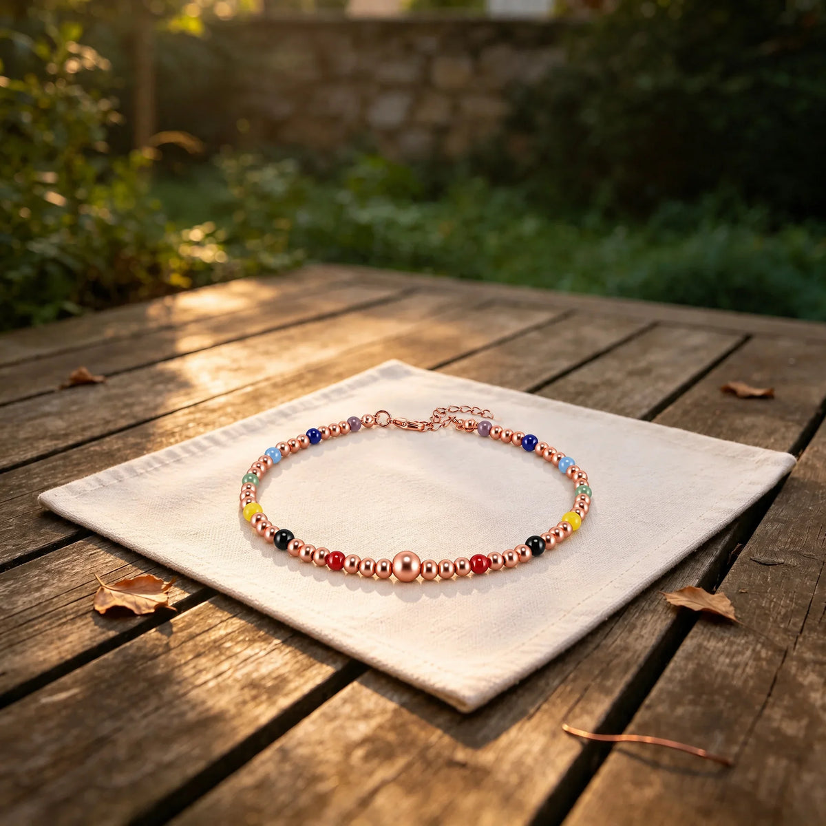 Copper bead anklet for women with seven colored chakra beads and an adjustable extension chain, resting on white linen cloth in a sunlit garden