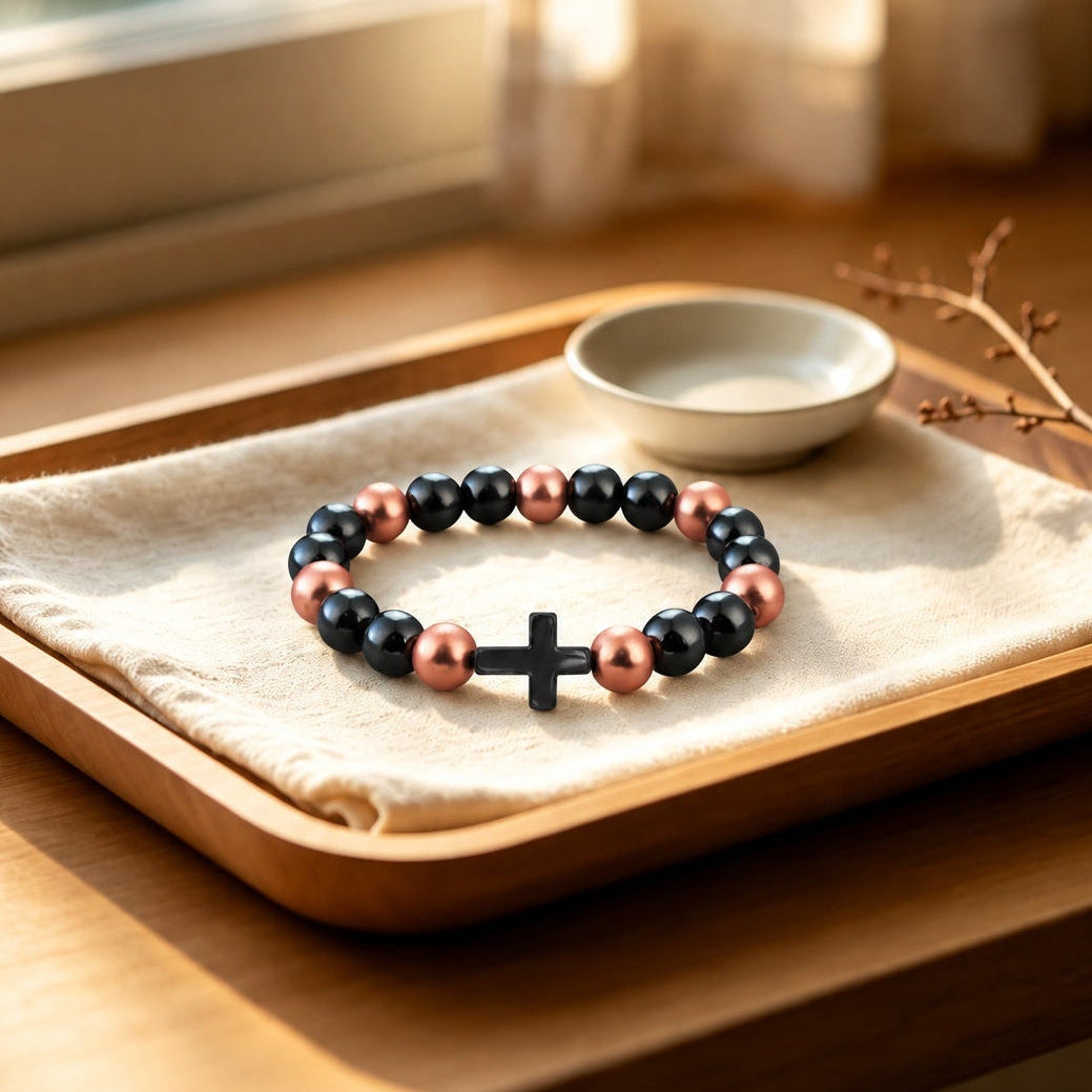 Magnetic copper bead bracelet with black cross pendant on wooden tray by window