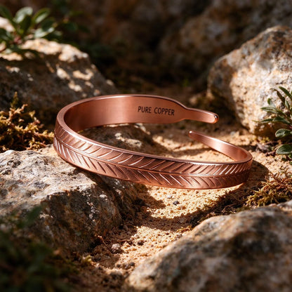   Pure copper leaf cuff bracelet with detailed feather pattern resting outdoors on natural rock and moss in sunlight.
  